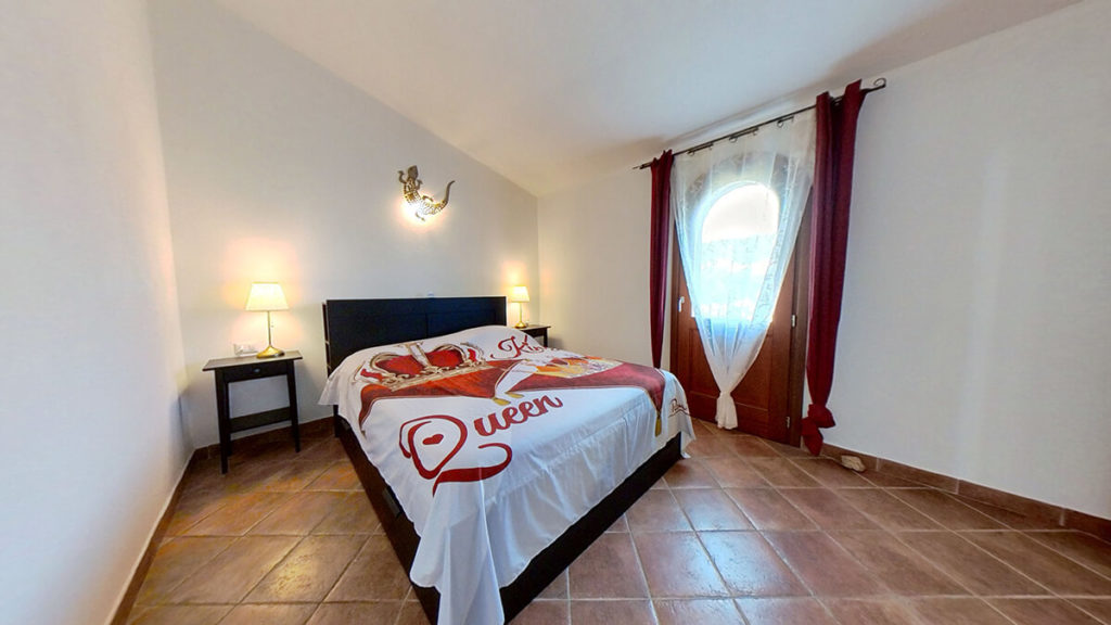 Stanza di albergo calda, con lenzuola King e Queen, e lampada a forma di geco sul muro. Tendaggio decorativo rosso sulla porta d'entrata.